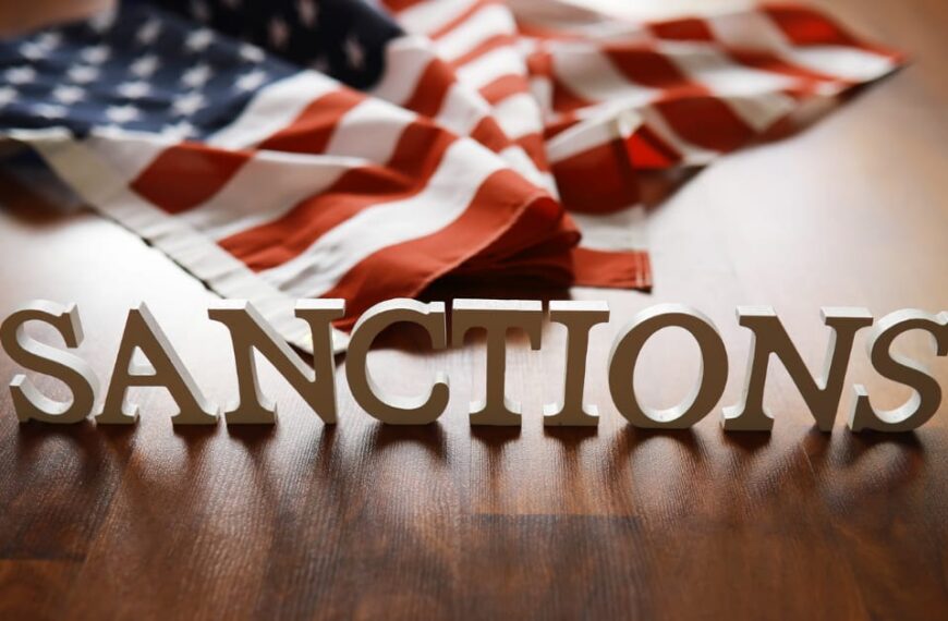 Economic Sanctions: Wooden letters spelling "SANCTIONS" placed in front of a folded U.S. flag on a wooden floor.