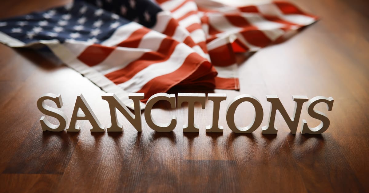 Economic Sanctions: Wooden letters spelling "SANCTIONS" placed in front of a folded U.S. flag on a wooden floor.
