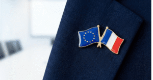 A close-up photograph of a lapel pin featuring crossed European Union and French flags on a blue suit jacket, symbolizing the European Nuclear Backstop partnership.