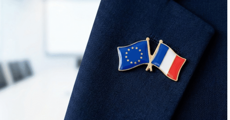 A close-up photograph of a lapel pin featuring crossed European Union and French flags on a blue suit jacket, symbolizing the European Nuclear Backstop partnership.